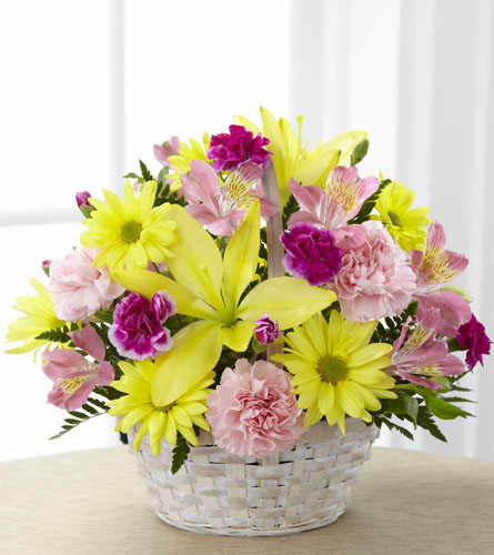 Floral arrangement of yellow and pink flowers in a white, woven handled basket.