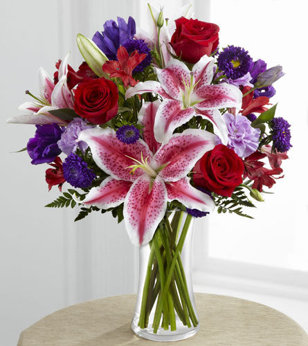 Arrangement of flowers with red roses, pink lilies, and purple flowers in a clear glass vase.