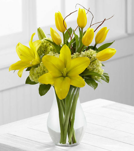 Bouquet of yellow lilies and tulips with greenery arranged in a clear glass vase.