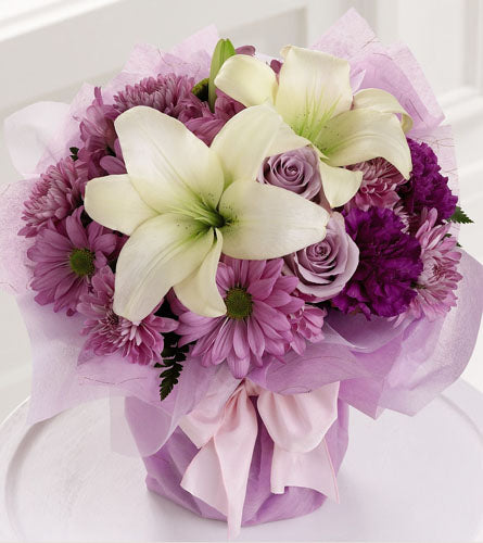 Pure Enchantment Bouquet - floral arrangement of white, purple and pink flowers in a purple wrapped vase with white bow.