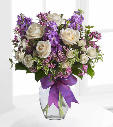 Enchanted Cottage - bouquet of white, purple and pink flowers in a clear vase with a purple bow.