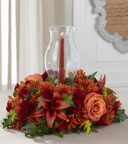 Heart of the Harvest Centrepiece - floral centrepiece of orange and burgundy flowers, arranged around a tapered candle with a hurricane glass holder.