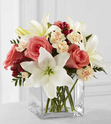 Arrangement of flowers with white lilies, coral roses, and pink mini carnations in a clear vase.