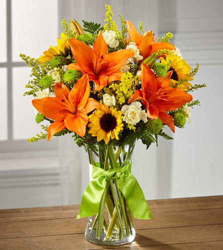 Country Calling Deluxe Bouquet - Arrangement of orange lilies, yellow sunflowers, and other mixed flowers in a clear vase tied with a green ribbon.