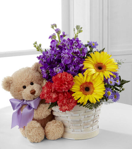 FTD's Best Year Bouquet - Teddy bear with a purple bow next to a white basket containing colourful flowers.
