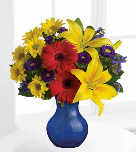 Summer Daydream Bouquet - yellow lilies, red gerberas, and purple asters arranged in a blue glass vase.